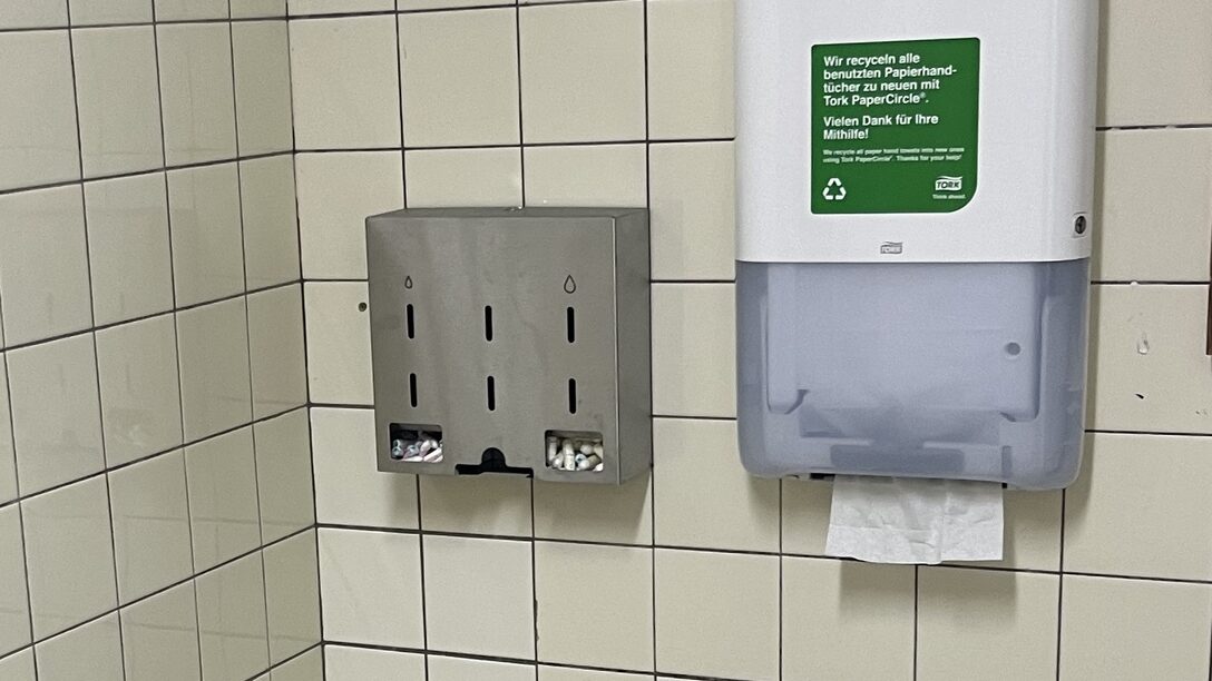 Ein Spender für Menstruationsprodukte hängt an einer gefliesten Wand. Daneben ein Handtuchspender.