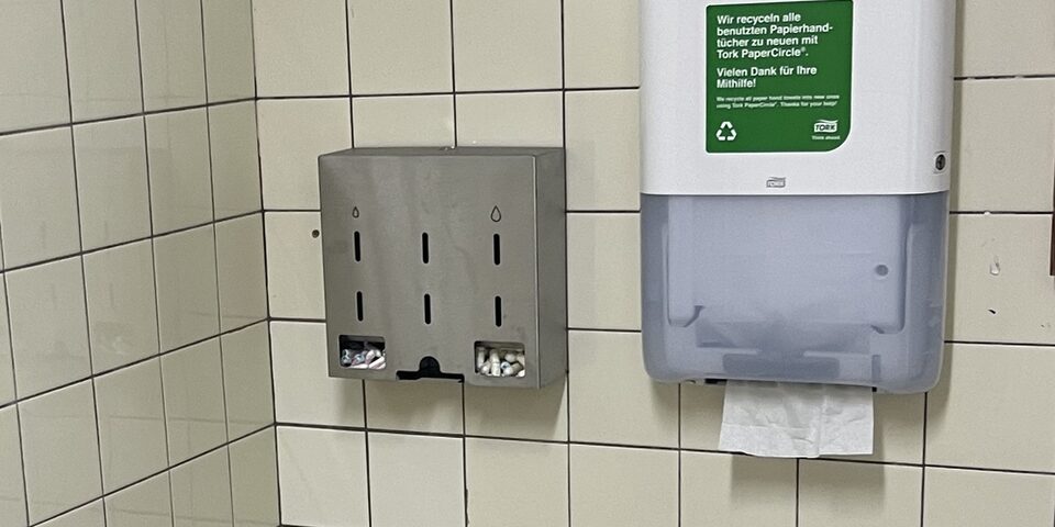 Ein Spender für Menstruationsprodukte hängt an einer gefliesten Wand. Daneben ein Handtuchspender.