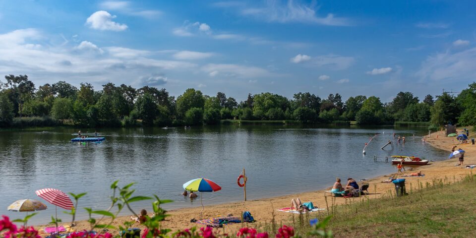 Sandstrand und Blick auf den See im Waldschwimmbad. Vereinzelt bunte Sonnenschirme und Liegeflächen.