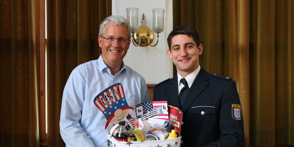 Oberbürgermeister Patrick Burghardt und Eduard Trippel halten einen Geschenkkorb mit Snacks aus den USA