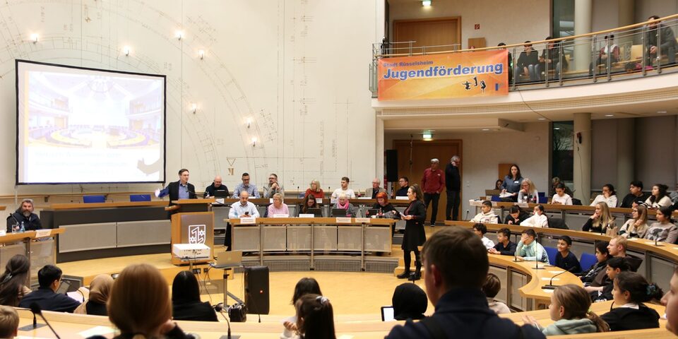 Bürgermeister Dennis Grieser steht am Rednerpult des mit Kindern und Jugendlichen gut gefüllten Ratssaals. An der Galerie hängt ein orangenes Banner mit der Aufschrift "Stadt Rüsselsheim. Jugendförderung".