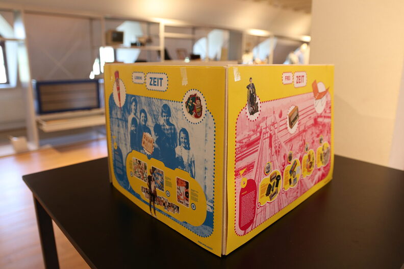 Modell des Pop-Up-Pavillons aus gelber Pappe steht auf einem schwarzen Tisch. Blaue und pinke Felder mit Fotos und Schrift sind als Aufdruck zu sehen. Im Hintergrund verschwommen die Regale der Sonderausstellung.