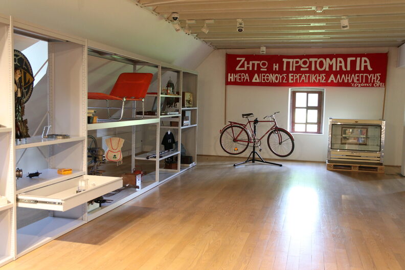 Blick auf die Regale der Sonderausstellung mit verschiedenen Exponaten. Daneben ein Fahrrad auf einer Halterung, darüber ein rotes Schrift-Banner mit griechischem Schriftzug.
