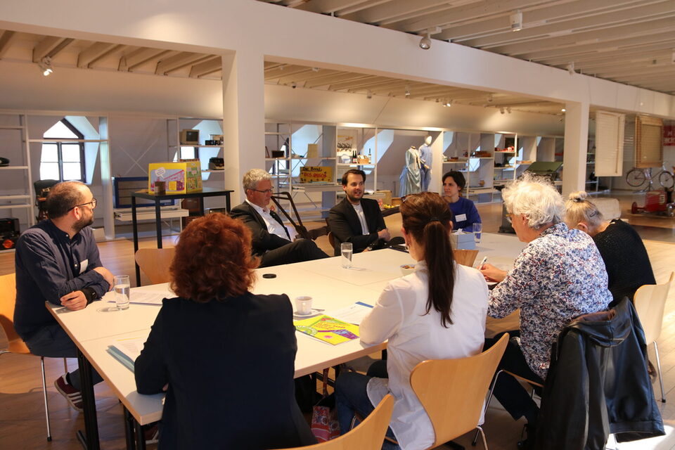 In den Räumlichkeiten der Sonderausstellung sitzen das Museums-Team, Oberbürgermeister Patrick Burghardt und Museumsleiter Nicolas Lange mit Journalistinnen und Journalisten an einem Tisch. Im Hintergrund die Regale des Schau-Depots.
