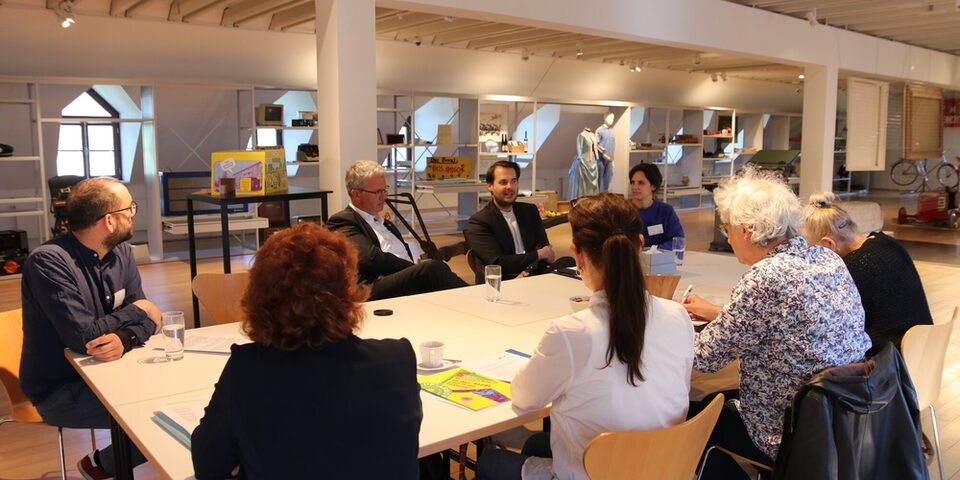 In den Räumlichkeiten der Sonderausstellung sitzen das Museums-Team, Oberbürgermeister Patrick Burghardt und Museumsleiter Nicolas Lange mit Journalistinnen und Journalisten an einem Tisch. Im Hintergrund die Regale des Schau-Depots.
