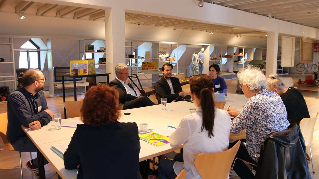In den Räumlichkeiten der Sonderausstellung sitzen das Museums-Team, Oberbürgermeister Patrick Burghardt und Museumsleiter Nicolas Lange mit Journalistinnen und Journalisten an einem Tisch. Im Hintergrund die Regale des Schau-Depots.