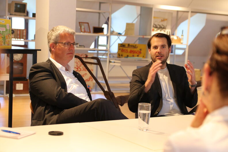 Nahaufnahme von Oberbürgermeister Patrick Burghardt und Museumsleiter Nicolas Lange, der gestikuliert. Beide sitzen an einem Tisch. Im Hintergrund die Regale des "Schau-Depots" in der Sonderausstellung des Museums.