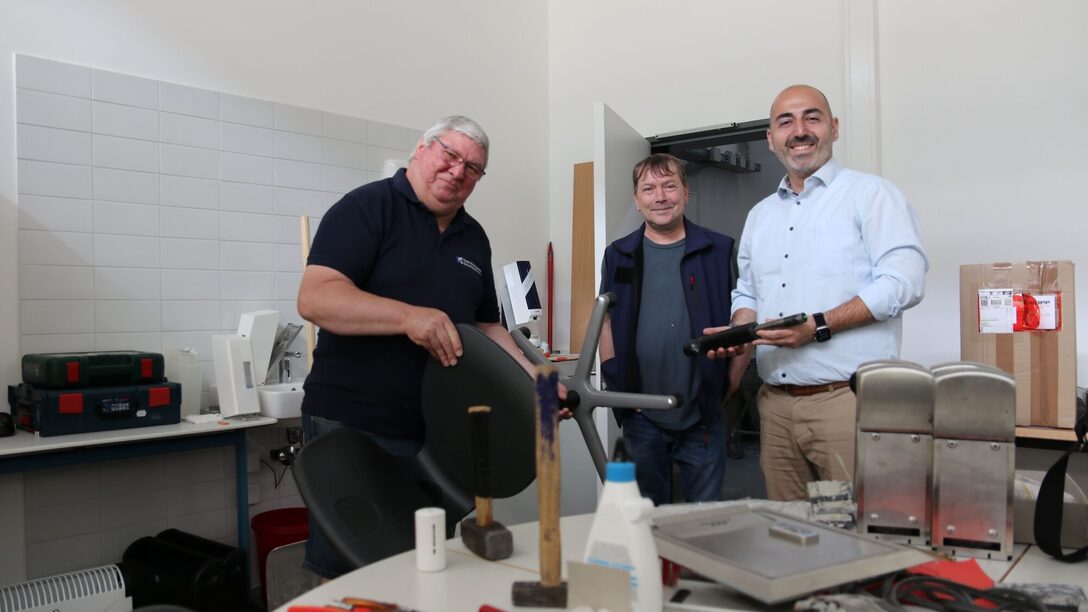 Hinter einem Tisch mit Werkzeug zeigen die Schulhausverwalter dem Bürgermeister einen Stuhl und die Druckluftfeder, die ausgetauscht werden musste.