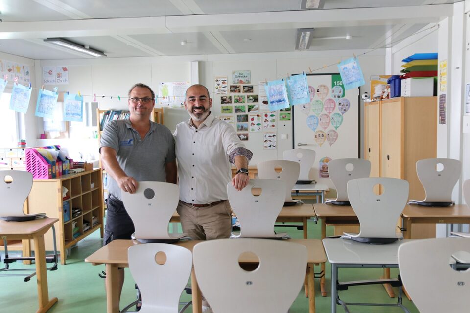 Der Schulhausverwalter der Eichgrundschule und der Bürgermeister stehen hinter einer Stuhlreihe in einem Klassenraum.