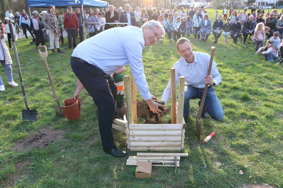 Im Vordergrund setzen Oberbürgermeister und Stadtrat den neuen Baum, im Hintergrund sieht man die Veranstaltung.