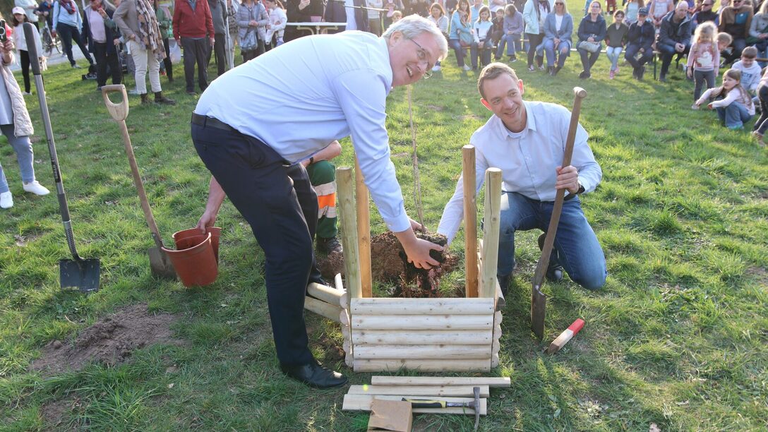 Im Vordergrund setzen Oberbürgermeister und Stadtrat den neuen Baum, im Hintergrund sieht man die Veranstaltung.