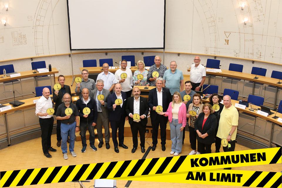Die Mitglieder des Haupt- und Finanzausschusses stehen vor der Leinwand im Ratssaal und halten gelbe Uhrschilder, deren Zeiger auf kurz vor 12 stehen, hoch. Das Foto ziert ein gelb-schwarz gestreiftes Banner mit der Aufschrift: Kommunen am Limit.