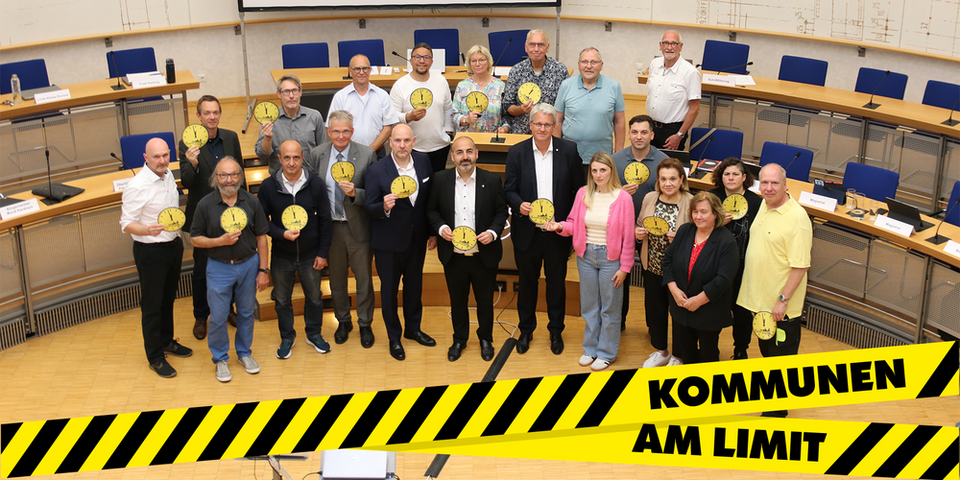 Die Mitglieder des Haupt- und Finanzausschusses stehen vor der Leinwand im Ratssaal und halten gelbe Uhrschilder, deren Zeiger auf kurz vor 12 stehen, hoch. Das Foto ziert ein gelb-schwarz gestreiftes Banner mit der Aufschrift: Kommunen am Limit.