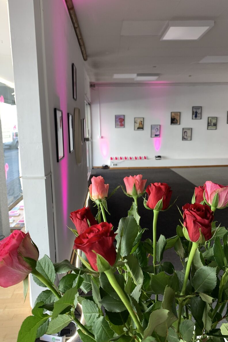 Ausstellungsraum freiraum f3. Im Vodergrund ist ein Strauß Rosen zu sehen, im Hintergrund hängen Bilder der Ausstellung an einer Wand.