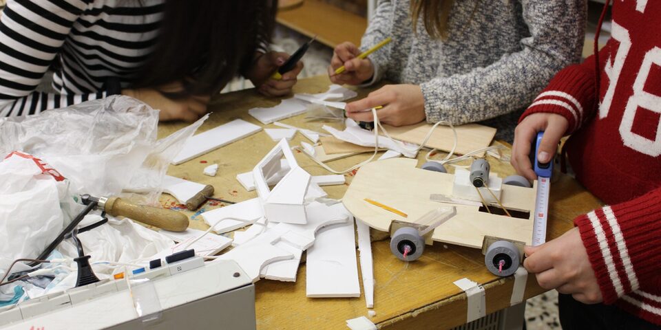 Drei Jugendliche bauen an einer Arbeitsplatte selbstgebaute Fahrzeuge aus Holz und Kunststoff zusammen.