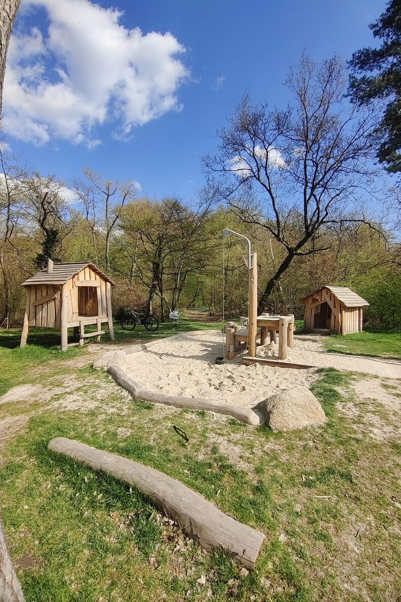 Im Vordergrund eine Sandspielfläche, im Hintergrund die neuen Spiel-Holzhütten des Räuberdorfs